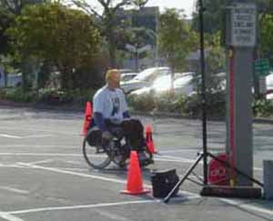 Figure 7: Photo. Manual wheelchair. A man is in a manual wheelchair in a parking lot. He is propelling himself by using his hands to turn push rims attached to the rear wheels.