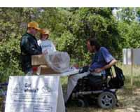 Figure 9: Photo. Power wheelchair. A man is in a power wheelchair. He is next to a table that has been set up alongside a trail.