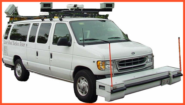 The photograph shows an extended white van equipped with four cameras mounted on support bars on the roof. Two cameras are on each side at the front of the van and two are at the rear of the van. A custom bumper the width of the van is mounted across the front of the vehicle parallel to the road. The piece of equipment contains lasers to capture pavement conditions and road geometry.