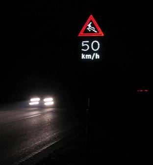 This picture is taken at night from the side of the road. It shows a vehicle approaching the photographer. At the top of the photograph is an illuminated sign which shows a deer (shown in white lights) jumping in the middle of a red-lighted triangle. Below the triangle is the speed limit 50 km/h (31.05 mi/h) illuminated in white.