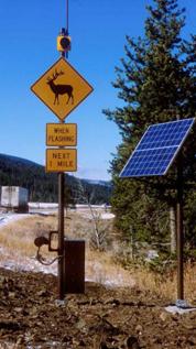 This picture shows two posts next to the roadway. The post furthest from the roadway has a solar panel on the top. The post closest to the roadway has a number of items attached to it. From top to bottom, this pole has an antenna, a flashing beacon, a yellow diamond sign with a silhouette of an elk, a yellow placard with the words WHEN FLASHING, a yellow placard with the words NEXT 1 MILE, a microwave transmitter, and a battery/controller cabinet.