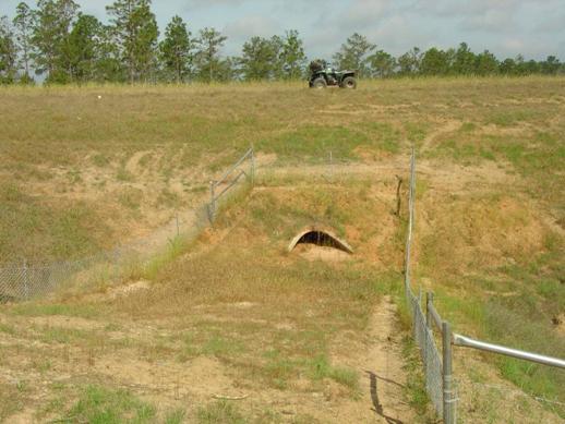 This is a picture of a small concrete arch culvert that goes under a roadway. Chain link fencing runs between the culvert and the roadway.