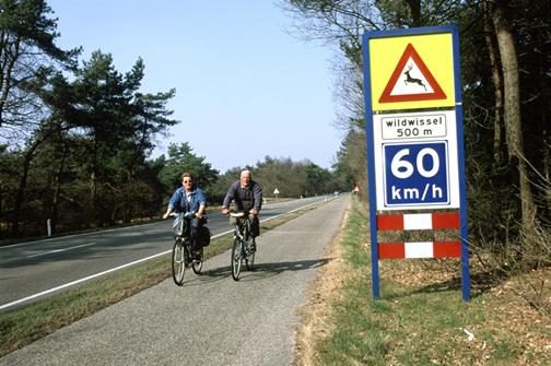  Figure 39. Photo. Advisory speed sign in The Netherlands located at a gap in exclusionary wildlife fencing (photo: Marcel Huijser). This picture shows a two-lane roadway with a paved pedestrian/bicycle trail next to it. Two bicyclists are riding toward the photographer and are passing a sign to their left. The top portion of the sign is a yellow rectangle, on which a white triangle with a red border is printed. Inside the white area of the triangle is a silhouette of a deer. The bottom portion of the sign is a 60 km/h speed limit sign (blue with white lettering).