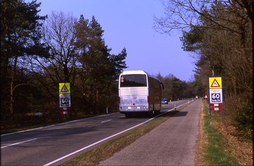 This is a picture of a roadway with two lanes. A large bus and a car drive toward each other. On either side of the roadway is an identical sign. The top portion of the sign is a yellow rectangle with a white triangle with a red border printed on it. Inside the white area of the triangle is a silhouette of a deer. The bottom portion of the sign is a 60 km/h speed limit sign (blue with white lettering). The fencing is not clearly visible in this picture.