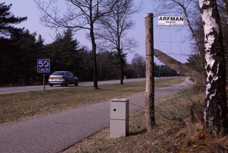 This picture shows the end of a fence line paralleling the roadway. At the end of the fence is a metal box that either transmits or receives an infrared, break-the-beam animal detection sensor. In the background the roadway can be seen with a speed limit sign for 50 km/h.