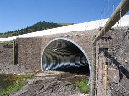 This is a picture of a large (roughly 2.4 m (8 ft) high) arch culvert going under a roadway. The culvert is corrugated metal with a sand and gravel layer in the bottom. Eight-foot deer-proof fencing that parallels the road can be seen ending on either side of the culvert.