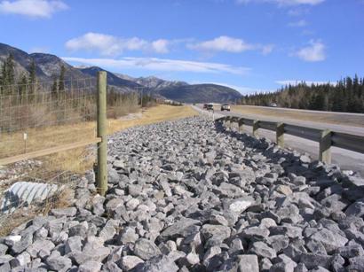 This is a picture of the end of a segment of deer-proof fencing. Between the fence end and the roadway, continuing beyond the fence end is a roughly 4.575-m (15-ft) wide swath of boulders (or rip-rap). The boulders average 0.305 m (1 ft) in diameter and go right up to the edge of the pavement. Most of the length of boulders are protected with guardrail that was probably already in place to protect a culvert seen in the lower left of the picture. The area of boulders beyond the guardrail had reflective delineators.