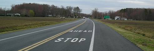 Figure 1. Photo. Example of a Rural STOP AHEAD Installation. The photo shows the words "STOP AHEAD" as pavement markings on a two-lane road in a rural area. They are painted in such a way that oncoming traffic sees the word "STOP" followed by the word "AHEAD," effectively making the marking say "AHEAD STOP" when read from top to bottom.