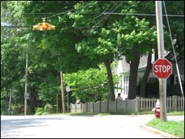 Figure 1. Photo. Example of Standard Overhead Flashing Beacon. A flashing beacon is suspended over the road in the center of a four-way intersection in a suburban area. There is also a STOP sign at the intersection. The beacon has four flashing lights so that one is facing in each direction.