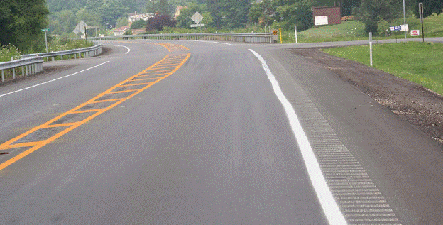 This figure provides an example of an implementation of concept 1. The photo was taken from the major approach at an intersection of 2 two-lane, two-way roadways with stop-control on the minor approach. There are paved shoulders with a white edge line and yellow center line. Concept 1 includes the introduction of a painted yellow median island on the major road approaches. The painted median begins as a double yellow centerline and gradually widens to a width of approximately 1.22 m (4 ft); the full median width is then continued to the intersection. Yellow cross-hatching is installed in the painted median. Rumble strips are also introduced on the outside shoulders and in the painted median on the major road approaches.