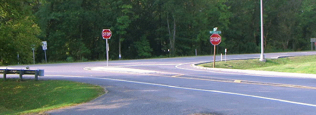 This figure provides an example of an implementation of concept 2. The photo was taken from the minor approach at a three-legged intersection. The major and minor roads are two-lane, two-way roadways with stop-control on the minor approach. The minor approach includes paved shoulders, white edgelines, a double yellow center line, and a STOP bar at the intersection with the major road. Concept 2 introduces a channelizing separator island on the minor approach, on which, a supplemental STOP sign is installed. The double yellow center line separates and a single yellow line continues around each side of the separator island; cross-hatching is installed between the yellow center lines where they split before the island. 