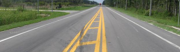 This figure shows an implementation of concept 1 at site 1 in Florida. The photo shows a section of the major road from a driver’s perspective looking away from the intersection. Shoulder rumble strips are shown to the outside of a white edge line, and the rumble strips end prior to the intersection. A painted median is shown with cross-hatching and median rumble strips that fully cover the width of the median between the inside edges of the pavement markings. The median rumble strips end prior to the intersection. A double solid yellow pavement marking is installed along each side of the median rumble strips, and raised pavement markings are installed along the centerline. 