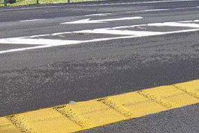 This figure shows an implementation of concept 1 at site 1 in Kentucky. The photo shows a section of the major road looking across the road from the centerline to the shoulder. The centerline is a solid yellow line, approximately 3.05 m (12 inches) wide and rumble strips cover the entire width. A portion of a left-turn lane is shown next to the center line, which is separated from the adjacent through lane by a white painted median with cross-hatching. The through lane has a white painted arrow, indicating the direction of travel and proper use of the through lane.