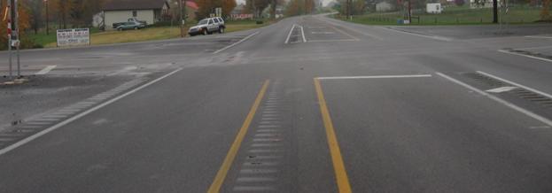 This figure shows an implementation of concept 1 at site 2 in Kentucky. The photo shows a section of the major road from a driver’s perspective approaching the intersection. Looking across the intersection, the photo shows a through and left-turn lane approaching the intersection and a through lane going away from the intersection in both directions. The left-turn lane has painted white arrows installed to indicate the direction of travel and proper use of the lane; STOP bars are also installed for the left-turn lanes. A painted yellow median, approximately 0.915 m (3 ft) wide, separates the left-turn and opposing through lane. The yellow median does not have cross-hatching. A white painted median, approximately 0.915 m (3 ft) wide, separates the left-turn lane from the adjacent through lane. The white median has cross-hatching installed. Rumble strips are installed along the shoulders and within the medians and extend to the intersection. In the painted medians, rumble strips are installed along left inside edge of the median that separates the left-turn lane from the opposing through lane and along the right edge of the painted median that separates the left-turn lane from the through lane in the same direction. Right-turn ramps with large turning radii are installed on both the major and minor roads.