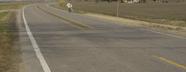 This figure shows an implementation of concept 1 at site 1 in Missouri. The photo shows a section of the major road looking away from the intersection. A painted median is implemented by splitting the double yellow center line and continuing the single yellow lines at a distance of approximately 4.06 m (16 inches) apart. Rumble stripes are installed to cover the entire width of the median, but no cross-hatching is present. Narrow paved shoulders, approximately 0.305 to 0.61 m (1 to 2 ft) wide, are present, and shoulder rumble stripes are installed along the shoulder. The painted median and all rumble stripes end approximately 15.25 m (50 ft) prior to the intersection. A single dashed yellow center line extends to the intersection.