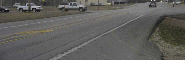 This figure shows an implementation of concept 1 at site 2 in Missouri. The photo shows a section of the major road looking toward the intersection with the minor road. A painted median is implemented by splitting the double yellow center line and continuing the single yellow lines at a distance of approximately 0.915 m (3 ft) apart. Cross-hatching is installed in the pained median, which extends to the intersection. Median rumble stripes nearly cover the entire width of median. A paved shoulder is present and is approximately 1.83 m (6 ft) wide. Shoulder rumble stripes are installed on the white edge line. All rumble stripes end 15.25 m (50 ft) prior to intersection.