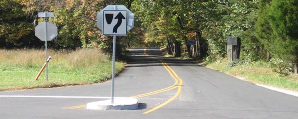 This figure shows an implementation of concept 2 at site 1 in Virginia. The photo shows one approach of the minor road looking from the intersection toward the minor road. The approach is two-lane, two-way, and divided by a double-yellow center line. There are no paved shoulders and no edge lines. A primary STOP sign is installed on the minor approach along with a STOP bar. A concrete separator island is installed on the minor approach at the intersection, and a secondary STOP sign is installed on the island. The yellow center line is continued along both sides of the separator island. The separator island is nontraversable with vertical edges. 