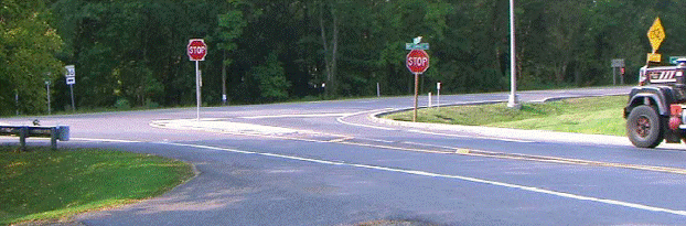This figure shows an implementation of concept 3 at site 1 in Maryland focusing on the minor approach. The photo shows the minor, stop-controlled approach of a three-legged intersection. The photo is taken from the left side of the minor road approach looking toward the intersection. The major and minor roads are two-lane, two-way roadways with stop-control only on the minor approach. The minor approach includes paved shoulders, white edge lines, a double yellow center line, and a STOP bar at the intersection with the major road. A separator island is installed on the minor approach, on which a supplemental STOP sign is installed. The double yellow center line separates prior to the island and a single yellow line continues around each side of the separator island; cross-hatching is installed between the single yellow lines prior to the island.