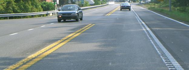 This figure shows an implementation of concept 3 at site 1 in Maryland focusing on the major approach. The photo shows a section of the major road from a driver’s perspective looking toward the intersection. The major road is two-lane, two-way with a double yellow center line, white edge lines, and approximately 2.44-m (8-ft) paved shoulders. There is no painted median in the application, but rumble stripes are installed on the center line and edge lines. A white dashed line is also shown across the minor road, connecting the white edge lines along the major road. Two vehicles appear in the photo traveling in opposite directions.