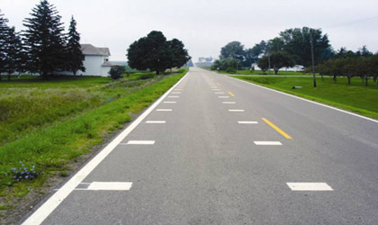 Photograph of a high-speed two-lane rural roadway just outside of Union, Iowa. The pavement is marked with transverse bars on the inside edges of the travel lane entering Union. The bars are spaced closer together as drivers approach the community.