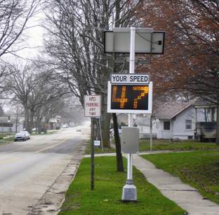 Photograph of an electronic speed feedback sign mounted on a post between the curb and sidewalk. The sign consists of a black-on-white panel with the legend "YOUR SPEED" mounted above a two-digit electronic sign displaying the number "47" by means of yellow lights on a black background. A solar panel is mounted at the top of the sign post. Homes can be seen on both sides of the road, as well as a few cars parked downstream on the opposite side of the road from the speed feedback sign.
