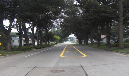 Photograph showing a main road through Union, Iowa, lined with large trees on both sides. There is a white edgeline painted on one side of the road about 8 ft from the curb. There is a painted center island in the middle of the road consisting of yellow striped markings around the outside and yellow cross-hatch markings in the center. 