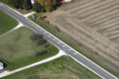 Aerial photograph showing white chevrons painted on the right side of the roadway in Roland, Iowa. The chevrons point in the direction of travel and are closer together and  thinner as drivers approach the community. Just downstream of the chevron markings is the number "25" painted on the black asphalt pavement using white markings followed by "MPH," also in white markings. There is a field on one side of the road.