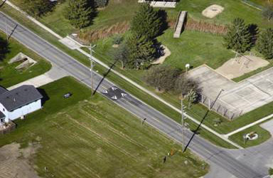 An aerial photograph showing a raised speed table placed across both lanes of travel in   Gilbert, Iowa. The ramps of the table have white chevron markings pointing in the direction of travel. A park area can be seen on the north side of the road.