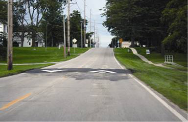 A photograph of the speed table from the driver viewpoint. Because it is only a few inches high, it almost appears flush with the roadway. The road appears to be on a slight grade, and there is a sidewalk on the right side of road that is setback about 15 ft from the edge of pavement.