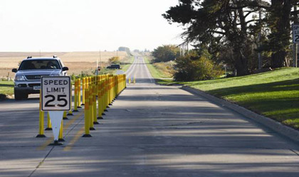 Photograph showing a center island consisting of two rows of  tall, yellow, tubular markers. Double yellow centerline markings run between the two rows of markers. A 25 mi/h speed limit sign is mounted close to the pavement on a flexible post at the nose of the island. The road is straight and the roadside environment just passed the island appears very rural