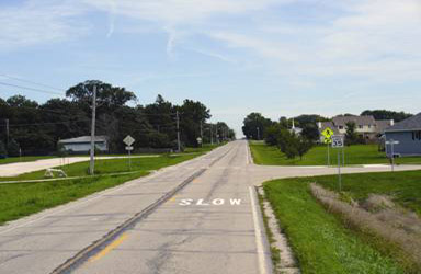 Photograph showing the word "SLOW" painted white in the center of the travel lane in  Slater,  Iowa.  The road has a grass shoulder, and a few houses can be seen in the background that are setback from the road. A 35 mi/h speed limit sign and a bright yellow-green pedestrian warning sign can be seen just past the "SLOW" marking.