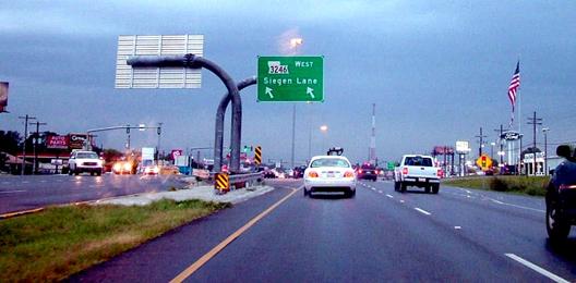 This photograph shows a large overhead sign above the left-turn lanes on the approach to the crossover at the Baton Rouge DLT. The sign is mounted on a large single-unit steel pole that is appears to be more than 3 m (12 inches) in diameter. The pole has a 90-degree bend. The sign has a route symbol for Mississippi State Route 3246 West. Below that symbol is the street name, Siegen Lane. Below the street name are two left slanted arrows, one centered over each of two left-turn lanes.