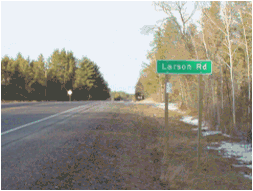 Figure 4. Photo. Example of an advance sign in Wisconsin. This photo shows an advance street name sign in Wisconsin, which is a rectangular green sign with a white border and white text. The sign is seen on the right shoulder of a long flat stretch of road. The sign is green with white text, reading "Larson Rd." The sign is mounted on two wooden posts. The intersection is not yet visible from this point.
