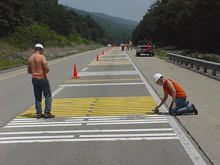 Figure 18.  Photograph.  Typical transverse test deck. The photograph illustrates yellow and white transverse pavement markings which lay across the right-hand lane of two lanes of one direction of a divided highway.  A road maintenance crew is working on the transverse pavement markings.  The crew members have placed orange cones along the lane as they work so that drivers do not enter the lane.