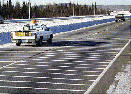 Figure 19.  Photograph.  Transverse test deck in Alaska.  The photograph illustrates white transverse pavement markings across two lanes in the same direction of a divided highway.  There is a white edge line on the right lane, and there are white dashed lines separating the two lanes.  Two cars are traveling in the left lane.