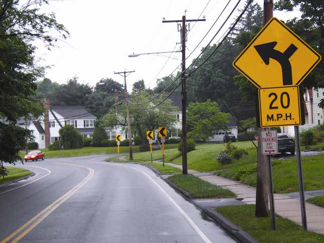 Figure 1. Photo. Example of curve treated with enhanced delineations in Connecticut. This photo shows an example of improved curve delineation along a two-lane undivided roadway with a curve to the left. The road is in a suburban neighborhood. There is a double yellow centerline and two white edge lines on the outside edge of the lanes, one in either direction, to designate the lanes. The roadway has curbs, gutters, and narrow shoulders. There are four utility poles shown along the outside of the curve on the right, and one pole has a street light attached. There is a narrow grass buffer on the right side of the road and then a sidewalk. Near the beginning of the curve, there is a curve warning sign. The sign is diamond-shaped and mounted into the ground with two posts. It is yellow with a black arrow that illustrates a 45-degree turn to the left, including a side road to the right along the curve. Below the sign, there is a supplemental plaque attached that reads, "20 M.P.H," indicating a 20-mi/h advisory speed. The supplemental plaque is square and yellow with black text. The following three signs in the sequence are chevrons, which indicate the presence and direction of the curve. These signs are yellow with the same black symbol that points to the left. They are mounted in the ground with one pole per sign. The black symbol is created by a black line from the upper right corner to the middle left of the sign and another black line from the middle left to the lower right corner, forming a point facing left. Each of the chevron sign posts also has a yellow reflective stripe. All curve warning signs include a "No Parking Anytime" sign on the sign posts.