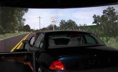 FHWA HDS. This photo depicts the Federal Highway Administrations (FHWA) Highway Driving Simulator (HDS). It shows the external rear view of a dark-colored automobile with a wrap-around projection screen in front of it. On the wrap-around screen, a daytime rural driving scene is portrayed with a roadway, pavement markings, a speed limit sign, grass, bushes, and trees.