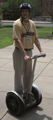 Figure 1. Photo. A rider on a SegwayTM HT. In this photo, there is an adult male standing on a SegwayTM Human Transporter (HT) on a sidewalk surrounded by grass. He is wearing a helmet, and his hands are placed in front of him on the handlebars.