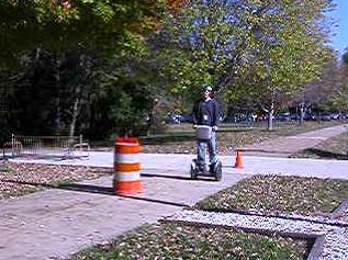 Figure 5. Photo. Wide section of the experimental sidewalk (without tape). The photo shows an adult male wearing a helmet and riding a SegwayTM Human Transporter forward on a wide section of the experimental sidewalk. Behind him, there is an orange cone in the center of two intersecting sidewalks. Directly in front of him, there is an orange- and white-striped barrel.
