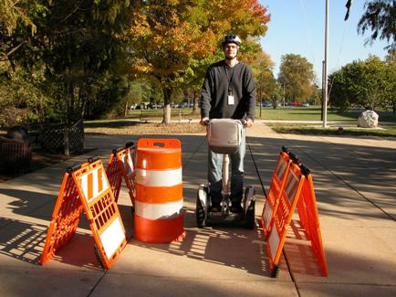 Figure 6. Photo. Narrow section of the experimental sidewalk (without tape). The photo shows an adult male wearing a helmet and riding a SegwayTM Human Transporter (HT) on a narrow section of the experimental sidewalk. He is photographed from the front and is moving forward. The narrow section is created from four orange barriers, two on each side of the SegwayTM HT rider, with an orange- and white-striped barrel between the four barriers but next to the two barriers on the left. He is riding on the right side of the barrel.