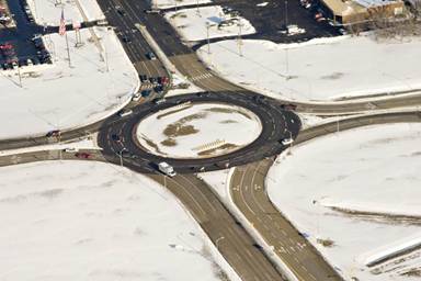 Figure 1. Photo. Michigan three-lane roundabout. This aerial photograph shows a three-lane roundabout at 18" Mile Road and Van Dyke Avenue in Sterling Heights, MI. The portion of Van Dyke Avenue that is visible in the photographs has six lanes, three in each direction. Van Dyke Avenue runs from the bottom right to the top left of the photograph. The roundabout entrances and exits for Van Dyke Avenue all have three lanes. The portions of 18½ Mile Road that are visible run horizontally across the photo and have four lanes, two in each direction. All entrances and exits that serve 18½ Mile Road have two lanes. The portions of the circular roadway that serve through movements for Van Dyke Avenue each have three lanes. The portions of the circular roadway that serve 18½ Mile Road through movements each have two lanes.