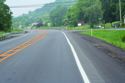 Figure 1. Photo. Lane narrowing using rumble strips at a rural Pennsylvania site. This photo shows a two-way road in a rural location. There are white edge lines to the right of the lanes in both directions. On the right side of the photo, rumble strips are on the right of the white edge line on the shoulder of the road. Between the two lanes, there is a painted yellow median island with yellow hatch markings. The rumble strips were also applied within the median island. This treatment narrows the smooth lane surface in both directions.