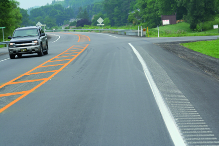 Figure 8. Photo. Cross hatching in median for better visibility of lane narrowing. This photo shows a two-way road, with one travel lane in each direction. Down the middle of the road, there is a yellow median with yellow hatching. On the right-hand shoulder, rumble strips are visible next to the edgeline. There is a motor vehicle driving forward in the left-hand lane.