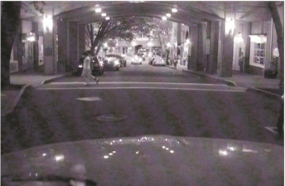 This photo shows a pedestrian crossing a street at an unsignalized crosswalk across the street from a vehicle in an urban environment.