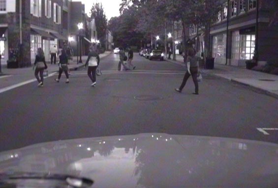 This photo shows people crossing a street at midblock in an urban environment in front of a vehicle. There are buildings on both sides of the street.