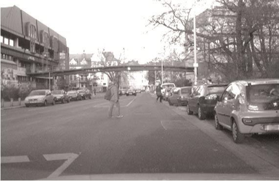 This photo shows pedestrians in an urban environment crossing a multilane street at midblock. There are parked vehicles on both sides of the street.