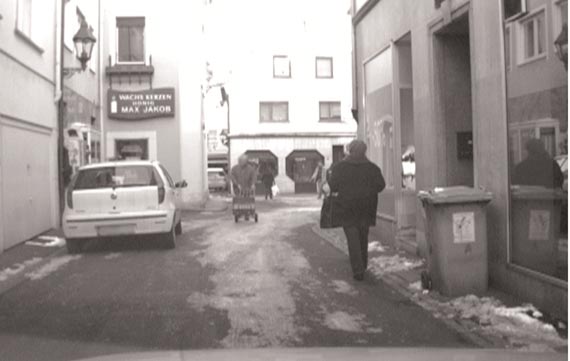 This photo shows people walking on a snow-covered street near a vehicle that is parked on the left side of the street.