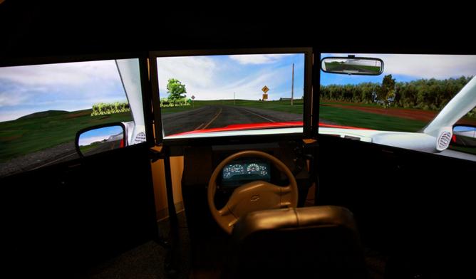 Figure 4. Photo. Driving simulator with three 42-inch plasma displays and driving buck. This photo shows the driving simulator used by study participants. A steering wheel and dashboard panel are in front of a chair, similar to the driver’s seat of a vehicle. Three flat-screen monitors are arranged around the chair, with one directly ahead and one at an angle on each side. The monitors show a simulated view out of the front and side windows of a vehicle along with rearview mirrors and the car frame. The screen images create the illusion of driving on a rural two-lane road. The simulator system is partially enclosed in a black tent so that the area around the monitors is mostly dark.