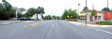 This photo shows 1 of 14 roadway panoramas presented to participants in the similarity rating task. Three lanes are visible in the direction of travel shown. On the right side of the roadway there is a sidewalk that directly abuts the roadway. A restaurant is on the right. A fluorescent warning sign can be seen next to newspaper racks on the sidewalk. There is a small median strip separating the three lanes in the direction of travel from the opposing lanes. There is a no U-turn sign in the median. On the left side of the highway, cars can be seen in a parking lot. There are trees in the distance.
