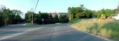 This photo shows 1 of 14 roadway panoramas presented to participants in the similarity rating task. The view is from the right lane of a four-lane undivided roadway that curves to the right in the distance. Buildings that appear to be four- or five-story apartments are directly ahead and visible above a line of small trees. To the right is a curb and unkempt grass that slopes upward away from the curb. A tractor and white truck are parked on the sloping grass. Telephone cables that stretch across the road originate behind the viewer on the right side of the roadway and cross to the left in the direction of the apartment buildings.